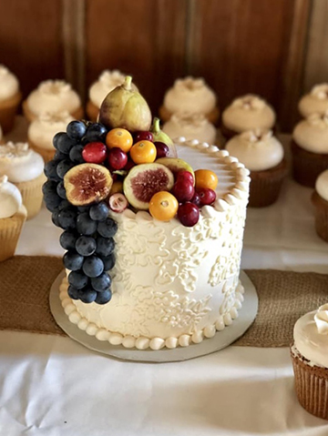 Small buttercream wedding cutting cake decorated with buttercream lace and fresh fruit, surrounded by cupcakes decorated with buttercream flowers.
