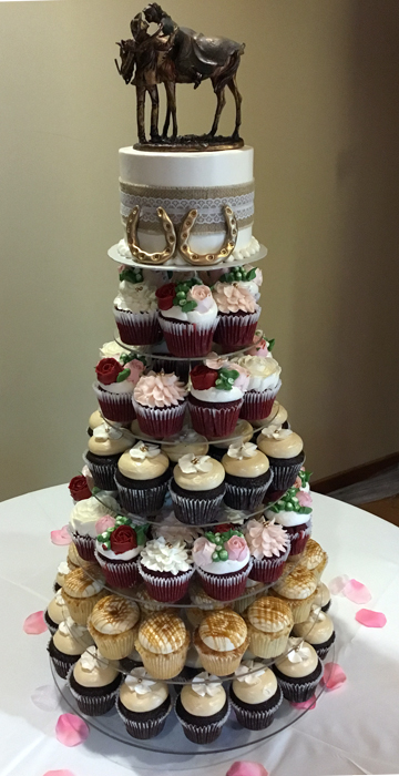 Small cutting wedding cake along with floral cupcakes and other decorated cupcakes displayed on round cake stand.