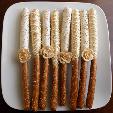 Tall pretzels sticks dipped in white chocolate with some painted gold and decorated with gold non perils, white chocolate stripes and gold sugar flowers