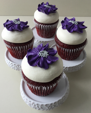 Red velvet cupcakes, topped with cream cheese icing and decorated with deep purple buttercream flowers with silver nonpareil centers